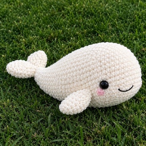 Puede incluir: Un peluche de ballena tejido a ganchillo de color crema con un cuerpo redondeado, una cola ancha y dos aletas laterales. La ballena tiene dos ojos de cuentas de plástico negro y pequeños círculos rosas bordados en las mejillas, con una sonrisa.