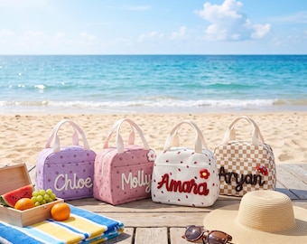 Puede incluir: Cuatro bolsas de almuerzo rectangulares con asas, cada una personalizada con los nombres Chloe en morado, Molly en rosa, Amara en blanco con fresas rojas y Aryan en un patrón de cuadros beige con un pequeño aplique de coche rojo.