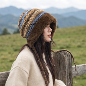 May include: A knit hat featuring a striped pattern in shades of brown, tan, and light blue, accented with a thick, dark brown faux fur trim around the face opening and long, braided side ties that hang down from the sides of the hat.