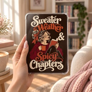 May include: A clear tablet case featuring a dark brown background with a graphic illustration of a person reading a book while holding a mug. The text reads "Sweater Weather & Spicy Chapters" in a stylised cream and orange font with a retro aesthetic.