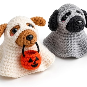 Puede incluir: Dos figuras de perro tejidas a ganchillo. Uno es un perro marrón con un disfraz de fantasma blanco que sostiene un pequeño cubo de calabaza naranja. El otro es un perro gris con orejas y hocico negros con un disfraz de fantasma gris.