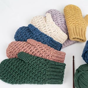 Könnte beinhalten: Eine Sammlung von sechs gehäkelten Fäustlingen in Dunkelgrün, Altrosa, Blau, Creme, Lavendel und Senfgelb, jeweils mit einem strukturierten Stichmuster und einem gerippten Bündchen, begleitet von passenden Garnknäueln und einer dünnen Häkelnadel.