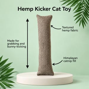 May include: A brown, cylindrical Hemp Kicker Cat Toy made of textured hemp fabric with Himalayan catnip fill. The toy is designed for grabbing and bunny-kicking, featuring a long, vertical shape with visible woven fiber details.