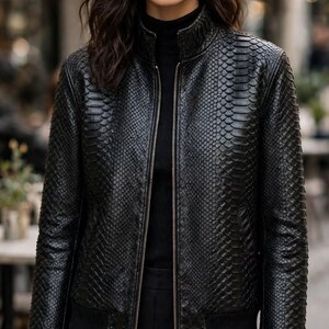 May include: A black bomber jacket featuring a textured snakeskin-like pattern, a high collar, and a full-length front zipper. The jacket has ribbed cuffs and a ribbed hem, worn over a black mock neck top and black trousers.