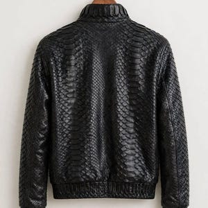 May include: A black jacket featuring a textured snakeskin pattern throughout the entire garment. The jacket includes a high collar, long sleeves with ribbed cuffs, and a ribbed hemline. The text leatheritemcrafts is printed at the bottom.