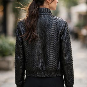May include: A black jacket featuring a textured snakeskin-like pattern throughout the exterior. The garment has a high collar, long sleeves with ribbed cuffs, and a ribbed hemline. The text leatheritemcrafts is visible at the bottom.