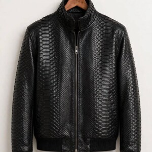 May include: A black jacket featuring a textured snakeskin pattern throughout the exterior. The garment includes a central silver-toned zipper, a high collar, and black ribbed cuffs and hem. The text leatheritemcrafts is visible at the bottom.