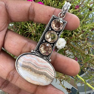 Puede incluir: Un colgante de plata con una gran piedra de ágata ovalada con bandas horizontales de color blanco, beige y rosa. Sobre la piedra ovalada, una barra vertical de plata sostiene tres piedras preciosas redondas, facetadas y de color marrón claro.