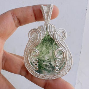 May include: A teardrop-shaped moss agate pendant encased in intricate silver-coloured wire wrapping. The stone features translucent white areas with deep green, fern-like inclusions. The metal wire forms decorative swirls and a woven border.