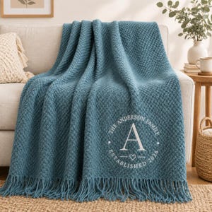 May include: A teal blue textured throw blanket with fringe edges, featuring a white circular monogram design that reads THE ANDERSON FAMILY, the letter A, a small heart icon, and the text ESTABLISHED 2026 printed on the lower right corner.