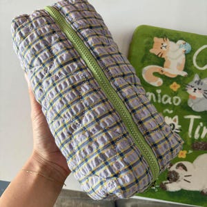Puede incluir: Un estuche acolchado con un patrón de cuadros en lavanda, amarillo y azul oscuro con una cremallera de color verde claro. La tela tiene una superficie texturizada y fruncida con un patrón de costuras repetitivo en la estructura rectangular.