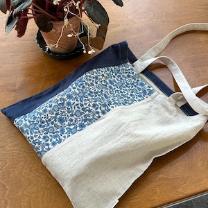 May include: A tote bag featuring a color-blocked design with a dark navy blue section, a central panel of blue and white floral print fabric, and a natural beige section. The bag includes two matching beige fabric handles and a small white label.
