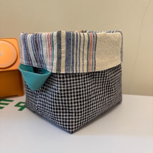 May include: A square fabric storage basket featuring a black and white gingham exterior pattern. The interior lining is made of a cream-coloured fabric with vertical blue, red, and green stripes. A small teal fabric handle is attached to the side.