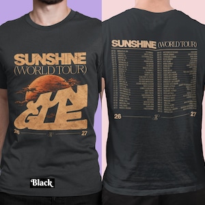 May include: A black short-sleeved t-shirt featuring a graphic print. The front displays the text SUNSHINE (WORLD TOUR) above a large orange sun and stylised lettering. The back lists tour dates and locations under the same tour title.