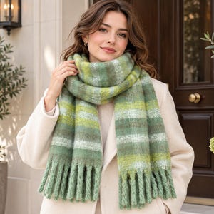 May include: A thick, fuzzy knit scarf featuring horizontal stripes in various shades of green, olive, and white. The scarf has a soft, textured appearance with long, twisted fringe detailing along the bottom edges, worn over a light beige coat.