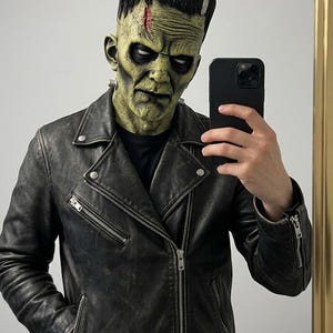 Op de afbeelding: Een Frankenstein-monstermasker voor het hele hoofd met een getextureerde groene huid, een zwart kapsel met platte bovenkant, een rode litteken op het voorhoofd en zilveren nekbouten, gedragen met een zwart leren motorjack met zilveren ritsen.
