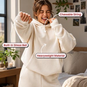 Könnte beinhalten: Ein cremefarbener Kapuzenpullover mit einer Kängurutasche auf der Vorderseite, einem integrierten Stressball und einer kaubaren Schnur. Das Kleidungsstück besteht aus schwerem Material und enthält Beschriftungen für diese Funktionen.