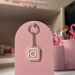 May include: A light pink keychain featuring the white Instagram logo icon. The square-shaped charm is attached to a silver-colored metal split ring by a short chain. The keychain is displayed hanging from a matching light pink arch-shaped stand.