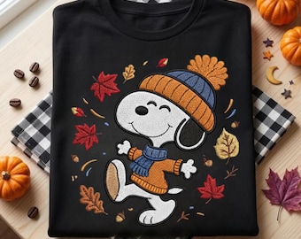 May include: A black crewneck t-shirt featuring a graphic of a white cartoon dog wearing an orange and blue knit hat and a matching orange sweater with a blue scarf. The design is surrounded by colourful autumn leaves and small acorns.