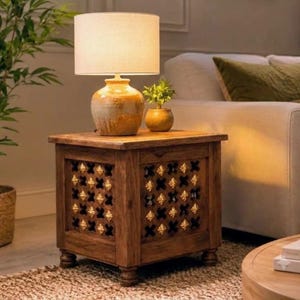 May include: A square wooden side table with four short legs and a lattice-patterned front panel featuring gold-colored accents. A ceramic lamp with a beige drum shade and a small potted plant sit on the flat top surface of the table.