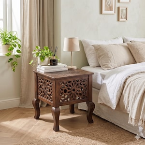 May include: A dark brown wooden side table featuring intricate geometric lattice cutouts on its sides and a carved floral motif on the square top. The table stands on four curved legs and supports a small lamp, two stacked books, and a potted plant.