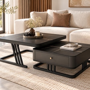 May include: A set of two square black coffee tables with gold-toned line accents and black metal frames. The lower table features a single drawer with a small gold knob, while the upper table holds a circular tray with a vase and candle.