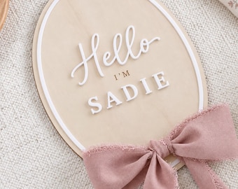 May include: An oval wooden plaque featuring a white raised border and the white 3D text Hello I'M SADIE. The plaque is decorated with a dusty rose-coloured fabric bow with frayed edges attached to the bottom, creating a soft and textured appearance.