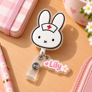 Può includere: Un porta badge retrattile con un coniglio bianco che indossa una cuffia da infermiere con una croce rossa. È dotato di un cinturino in plastica trasparente con bottone a pressione in metallo e un ciondolo in acrilico con il nome Lily in rosa.