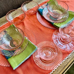 May include: A collection of vintage pink glass teacups, saucers, and bowls arranged on a coral-colored tablecloth. The glassware has a ribbed design. Green and patterned napkins add color to the table setting.