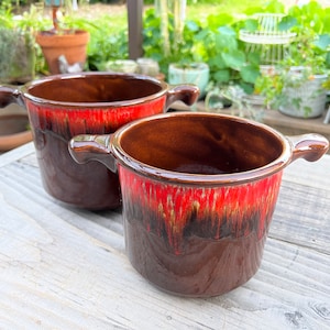 May include: Two brown ceramic pots with red and black glaze. The pots have two handles on each side and are sitting on a wooden surface.