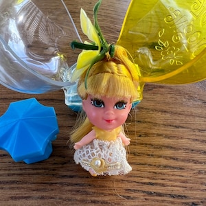 Könnte beinhalten: Eine Vintage-Puppe mit blondem Haar, blauen Augen und einem gelben Kleid mit Spitzenrock. Die Puppe hat eine Blume im Haar und befindet sich neben einem blauen schirmförmigen Behälter und einem transparenten Kunststoffbehälter mit gelben Flügeln.