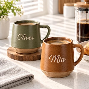 Puede incluir: Dos tazas de cerámica con acabado moteado, una de color verde oliva con el nombre Oliver y otra de color marrón ámbar con el nombre Mia. Cada taza cuenta con un asa redondeada y una base de color tostado natural con estrías.