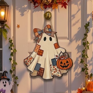 May include: A hanging wooden door sign shaped like a ghost with a patchwork pattern, a plaid bow, and a pumpkin bucket. The ghost features black eyes and is decorated with various autumn-themed fabric prints in orange, blue, and brown tones.