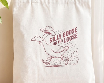 Pode incluir: Uma sacola de lona branca com um gráfico bordô de um ganso usando um chapéu de cowboy e botas, correndo com uma trilha de poeira atrás. O texto "SILLY GOOSE ON THE LOOSE" está impresso em bordô ao lado da ilustração do ganso.