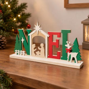 Op de afbeelding: Een houten tafeldecoratie die het woord NOEL vormt in groene en rode letters. De letter O is vervangen door een beige kerststalscène onder een ster. De decoratie bevat groene dennenbomen, een wit hekje en een wit rendier met een rode strik.
