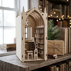 May include: A light-colored wooden book nook insert featuring a gothic-style arched structure. The interior displays a miniature library with shelves filled with books, a small desk, a chair, a ladder, and a staircase, all crafted from laser-cut wood panels.