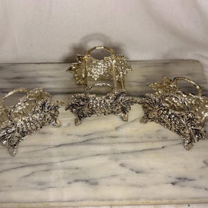 May include: Three silver-toned metal napkin rings featuring a detailed embossed design of grape clusters and vine leaves. Each ring has a rounded top handle and a sturdy base, showcasing a metallic finish with intricate textures.