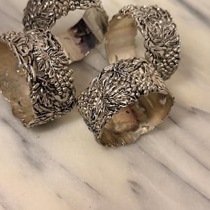 May include: A set of four silver-toned metal napkin rings, each featuring a detailed, raised relief pattern of grape clusters and vine leaves. The rings have a scalloped edge design and a polished, reflective finish throughout the surface.
