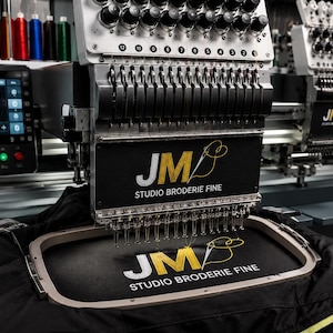 Peut inclure: Une machine à broder industrielle Tajima avec de multiples aiguilles et des bobines de fil de diverses couleurs. La machine dispose d'un écran numérique et d'un cadre noir affichant le logo blanc et jaune JM STUDIO BRODERIE FINE.