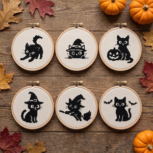May include: Six circular wooden embroidery hoops containing white fabric with black cross-stitch designs of cats. The designs feature a stretching cat, a cat in a witch hat, a cat with a jack-o'-lantern, a cat in a witch hat, a cat on a broom, and a cat with bats.