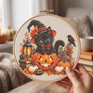 May include: A circular wooden embroidery hoop containing a cross-stitch pattern of a black cat wearing a witch hat and a plaid bow tie, sitting on a carved jack-o'-lantern. The scene includes autumn leaves, a glowing lantern, and a small bird.