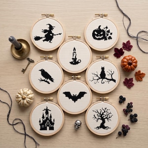 May include: Eight circular wooden embroidery hoops containing white fabric with black cross-stitch designs featuring Halloween motifs including a witch, jack-o'-lantern, candle, raven, cat, bat, haunted house, and a bare tree.
