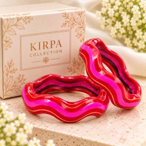 May include: Two wavy bangles featuring alternating bands of bright red and hot pink with thin gold-tone accents. The bracelets are positioned in front of a beige box with the text KIRPA COLLECTION printed in brown with floral illustrations.