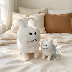 以下が含まれることがあります： 黒い刺繍の目と口を持つ、様式化されたウサギの形をした2つの白いぬいぐるみ。大きい方のウサギは直立しており、小さい方のウサギは背中に水色のループが付いており、機能的なアクセサリーとして機能しています。
