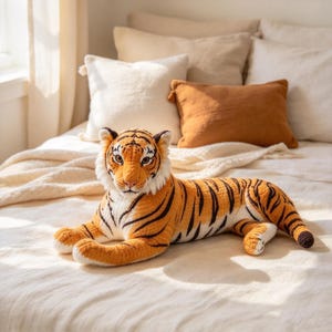May include: A plush tiger toy featuring a textured orange and white body with black stripes. The stuffed animal is depicted in a lying position with its front paws extended forward, showcasing detailed facial features including eyes and ears.