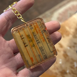 May include: A clear, rectangular keychain with a gold chain. The keychain has a vintage-style design with text that reads "RS MAMIE DRURY" and "RFD 2". The background is a light brown color.