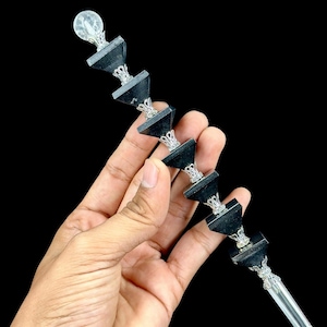 Könnte beinhalten: Ein dekorativer Stab mit sechs schwarzen, pyramidenförmigen Perlen, die durch verzierte silberfarbene Metallzwischenstücke getrennt sind. Die Spitze ist mit einer klaren, kugelförmigen Kristallkugel versehen, das untere Ende mit einer klaren Kristallspitze.