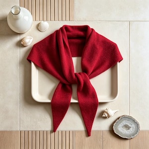 May include: A vibrant red knitted scarf with a triangular shape and pointed ends, featuring a soft, textured fabric construction. The scarf is draped in a central knot, showcasing its flexible material and consistent deep red colour throughout.