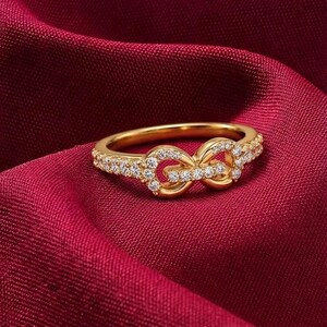 Op de afbeelding: Een gouden ring met een centraal oneindigheidssymbool versierd met kleine, heldere, fonkelende steentjes. De ringband is aan de zijkanten ingelegd met een rij bijpassende heldere steentjes, wat zorgt voor een glanzende en verfijnde uitstraling.