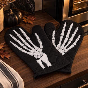 May include: A pair of black quilted oven gloves featuring a white skeletal hand print on the front. The design displays the bones of the fingers, palm, and wrist in a high-contrast graphic style. The fabric has a diamond-stitched pattern.